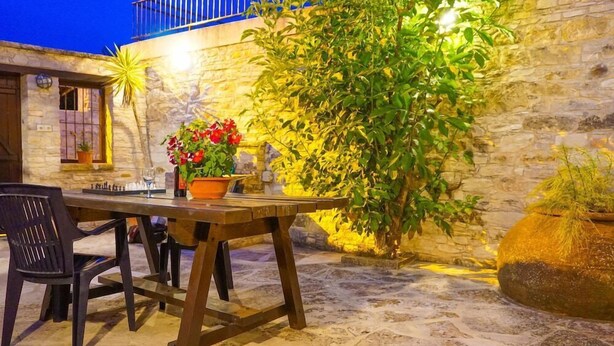 Une terrasse extérieure avec une table et des chaises en pierre, entourée de plantes en pot et d’un mur en pierre.