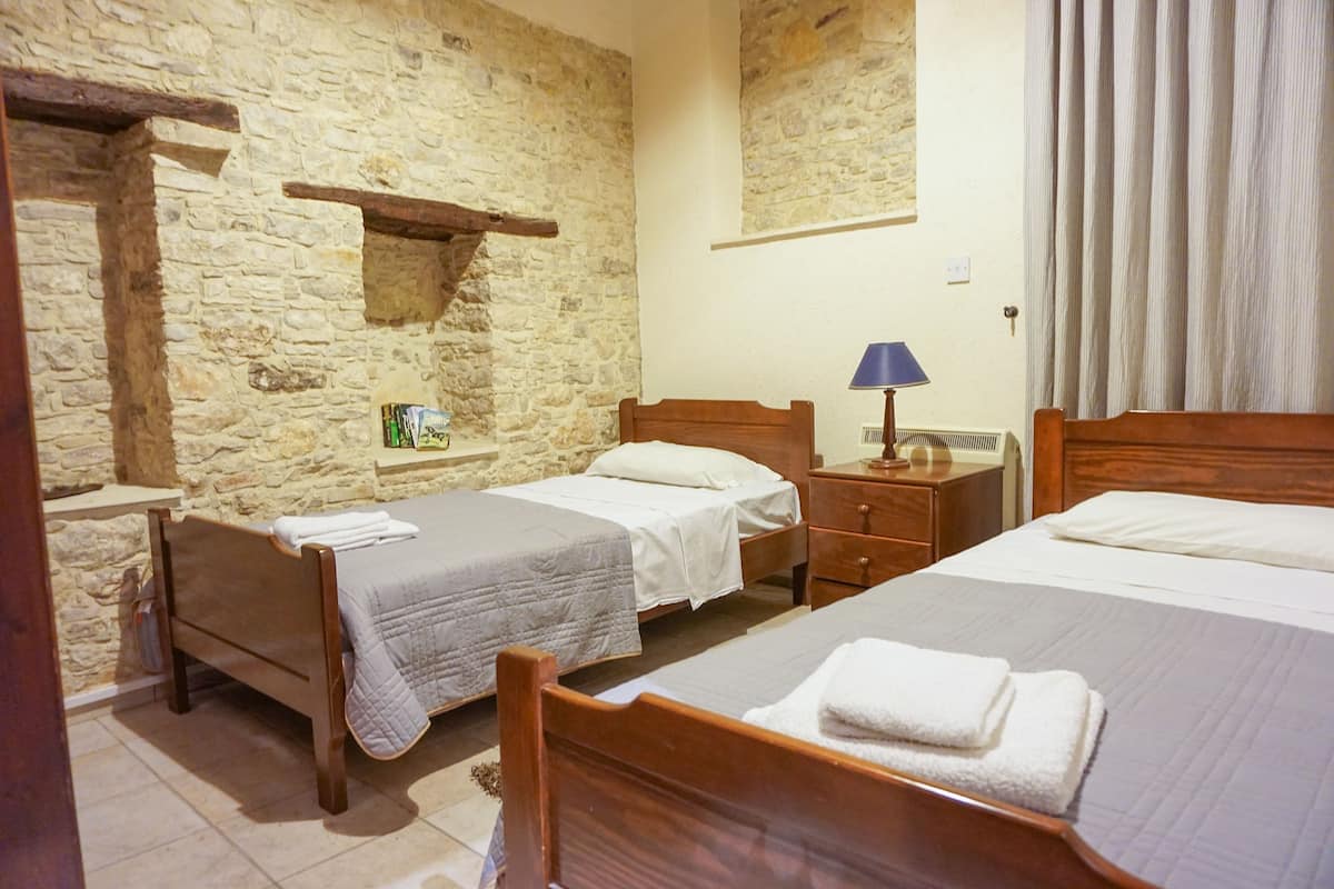 Quarto com duas camas de madeira, uma parede de pedra, um pequeno prateleira, uma mesa de cabeceira e um abajur.