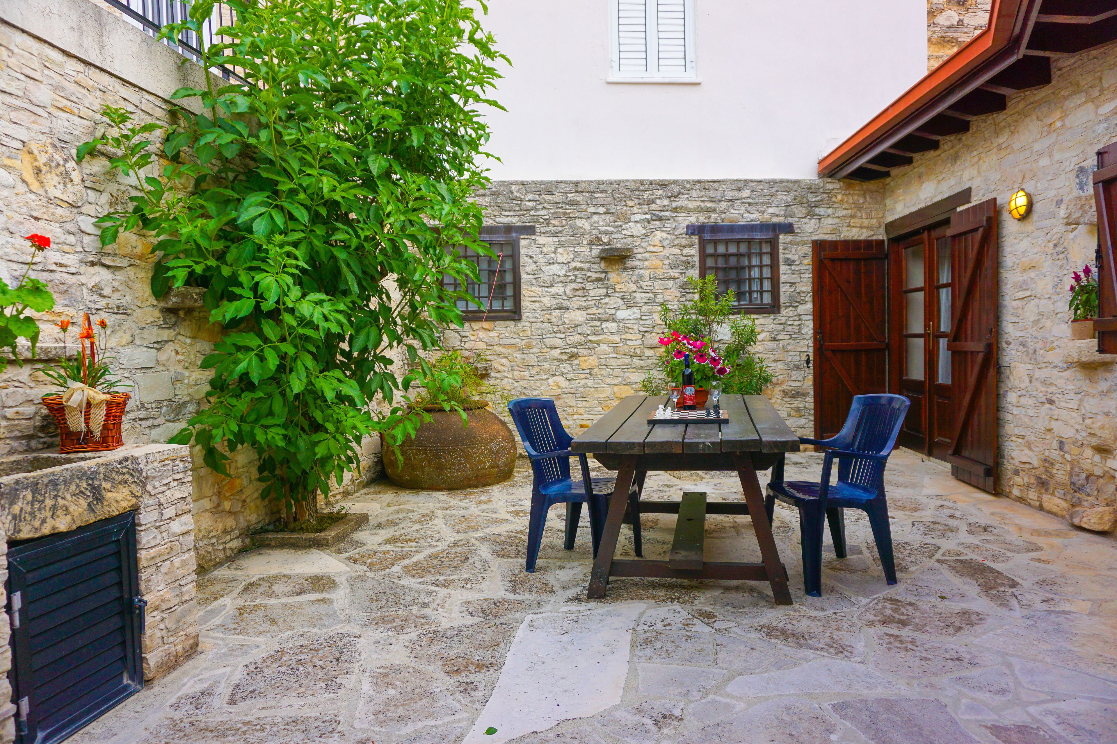 A stone courtyard with a wooden table and blue chairs, surrounded by stone walls and potted plants.