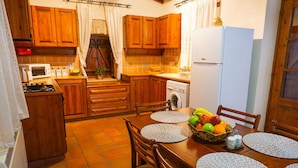 A kitchen with wooden cabinets, a stove, a microwave, a washing machine, and a dining table with placemats and a fruit basket. - Omodos Village Houses (Omodos)