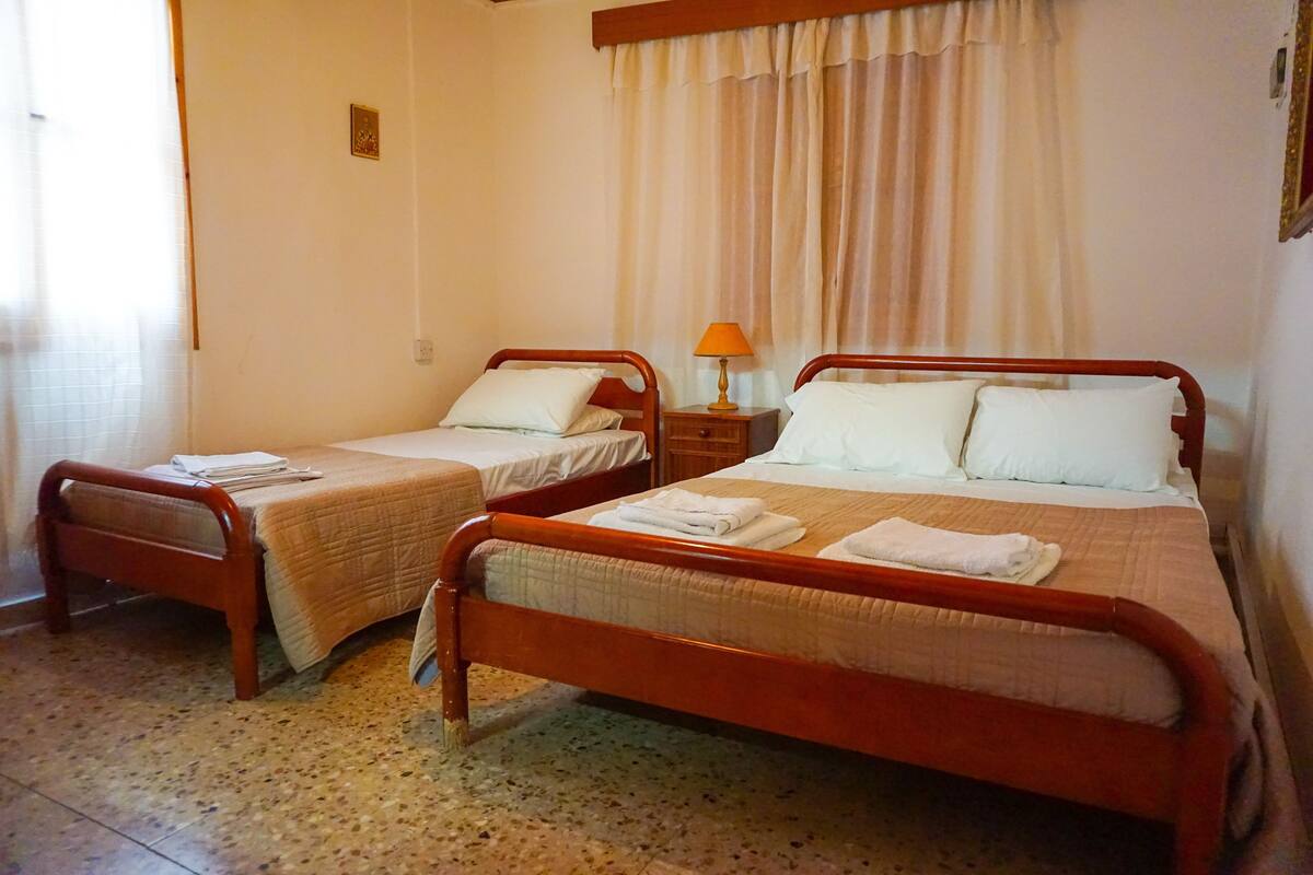Quarto de hotel com duas camas de solteiro, mesa de cabeceira com abajur e janela com cortinas.