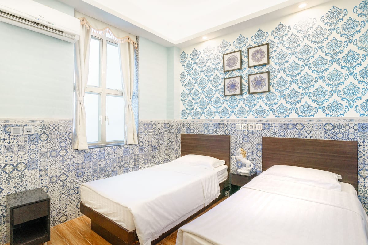 A room with two beds, a window with curtains, a wall with patterned tiles, and a nightstand with a lamp.