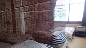 A room with a bed covered in striped bedding, a bedside table with a heater, and a window with blinds. - Guest House Shikotsu Kamui - Hostel (Chitose)