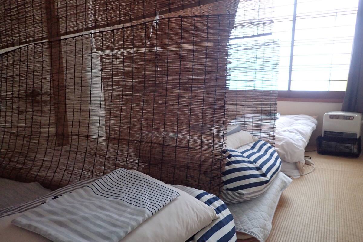 Une chambre avec un lit recouvert d’une couette à rayures, une table de chevet avec un radiateur et une fenêtre avec des stores.