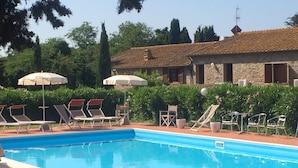 A swimming pool with lounge chairs and umbrellas, surrounded by greenery and a stone building.