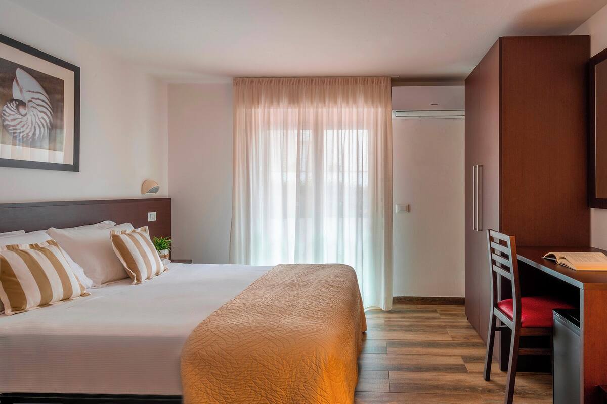 Una habitación de hotel con una cama, un escritorio, una silla y un ventanal con cortinas.