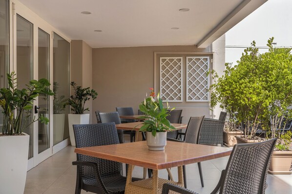 Un patio con mesa de madera y sillas de mimbre, rodeado de plantas en macetas y vistas a un edificio con grandes ventanales.