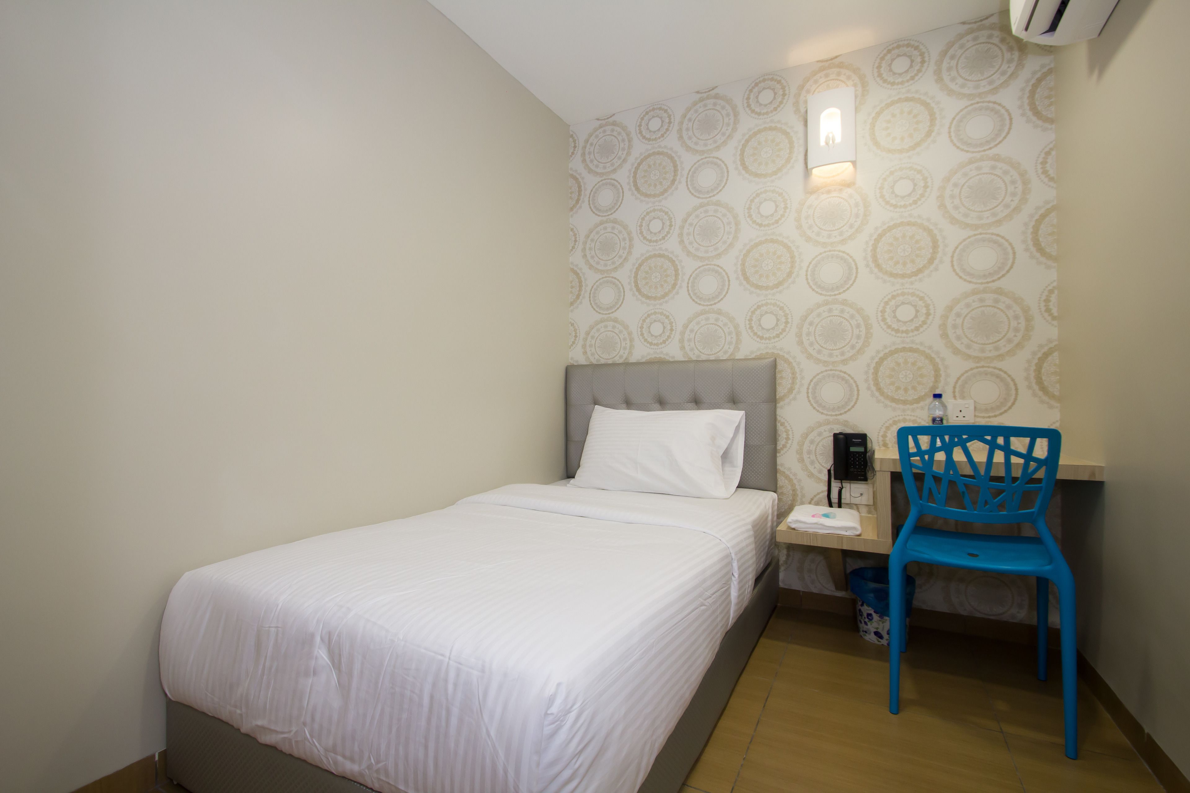 A small, neatly arranged bedroom with a single bed, a chair, and a desk with a telephone.