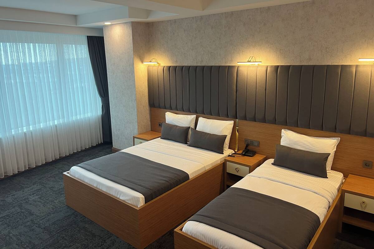 A hotel room with two beds, a large window with curtains, a headboard with built-in lighting, and a carpeted floor.