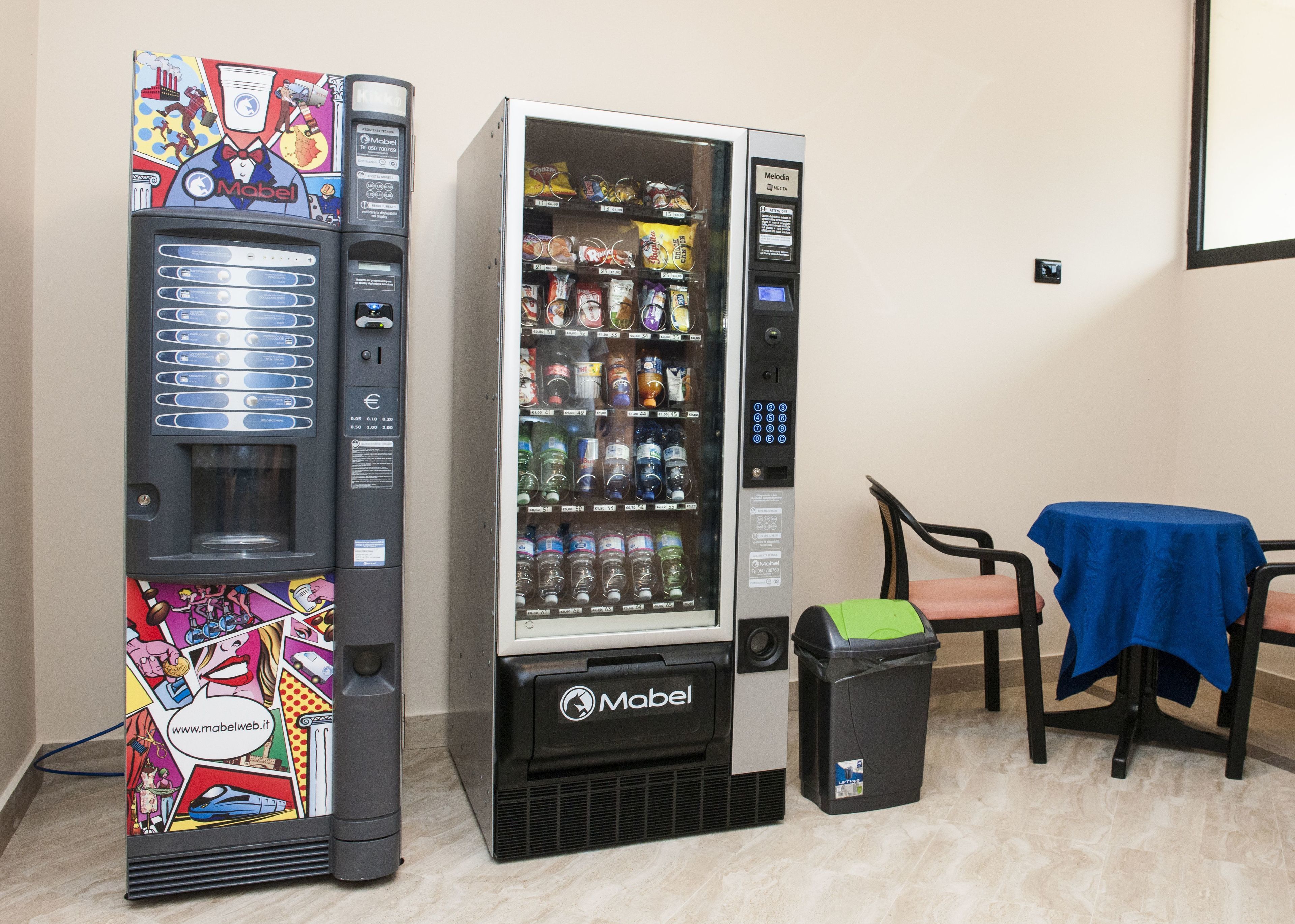Two vending machines with a variety of products, a trash bin, and a table with a blue tablecloth.