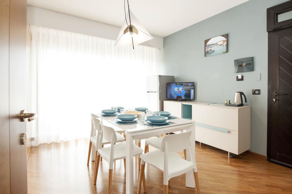 A dining area with a table set for four, white chairs, a modern light fixture, a television on a cabinet, and a door with a handle.