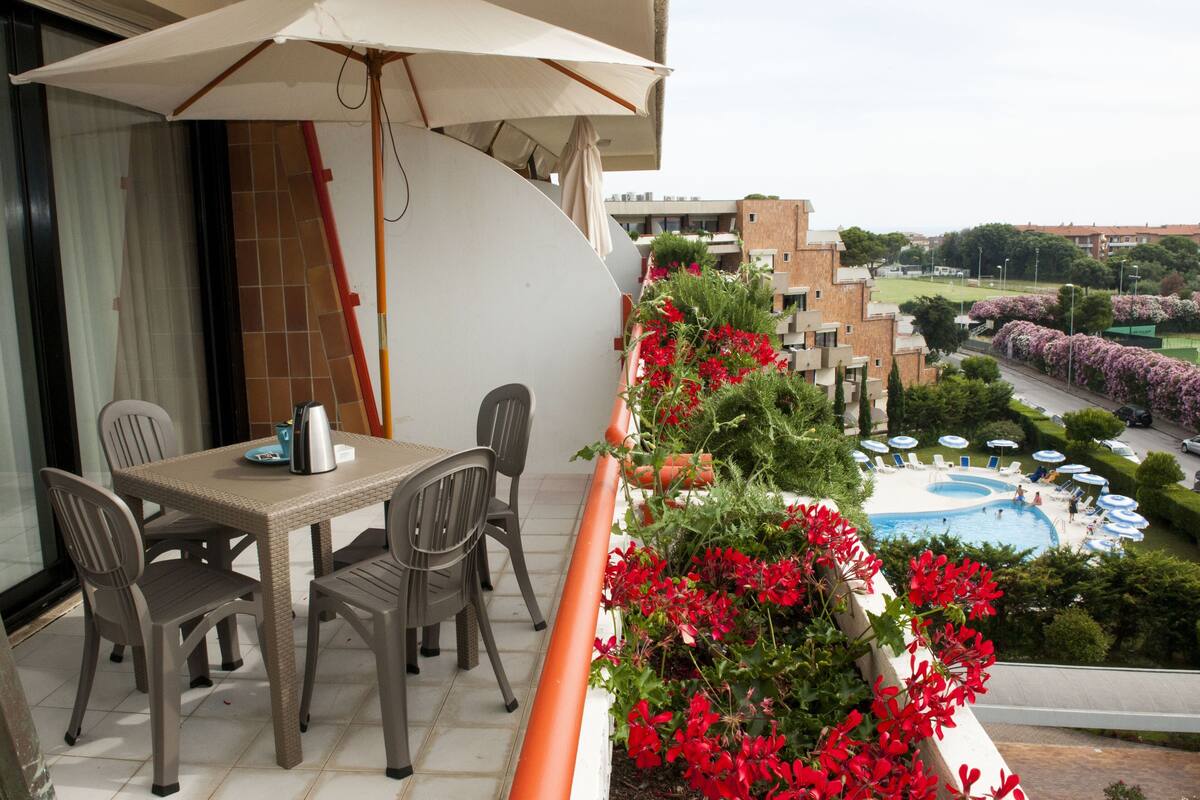 A balcony with a table and chairs, a pool area with umbrellas, and a view of surrounding buildings.