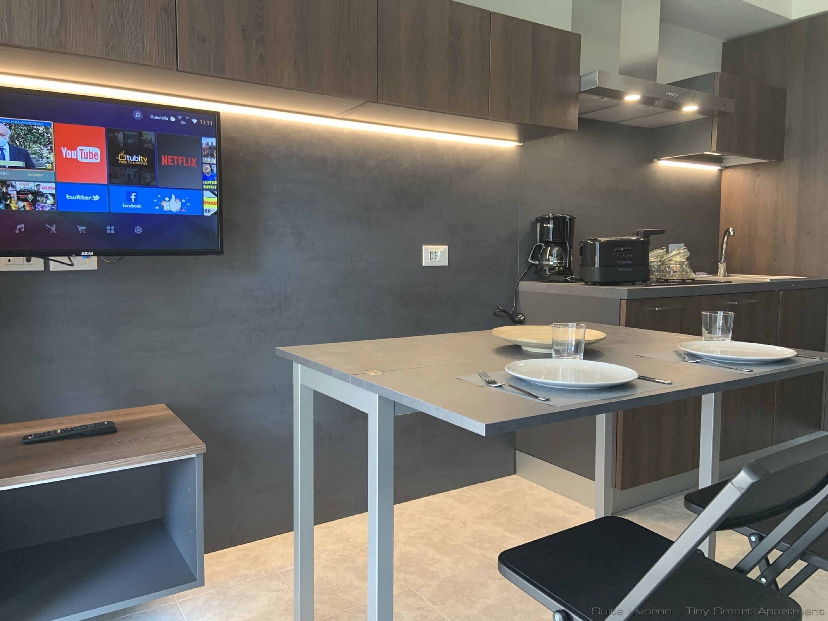 A modern kitchen with a flat-screen TV displaying various streaming services, a coffee maker, and a dining area with a table and chairs.