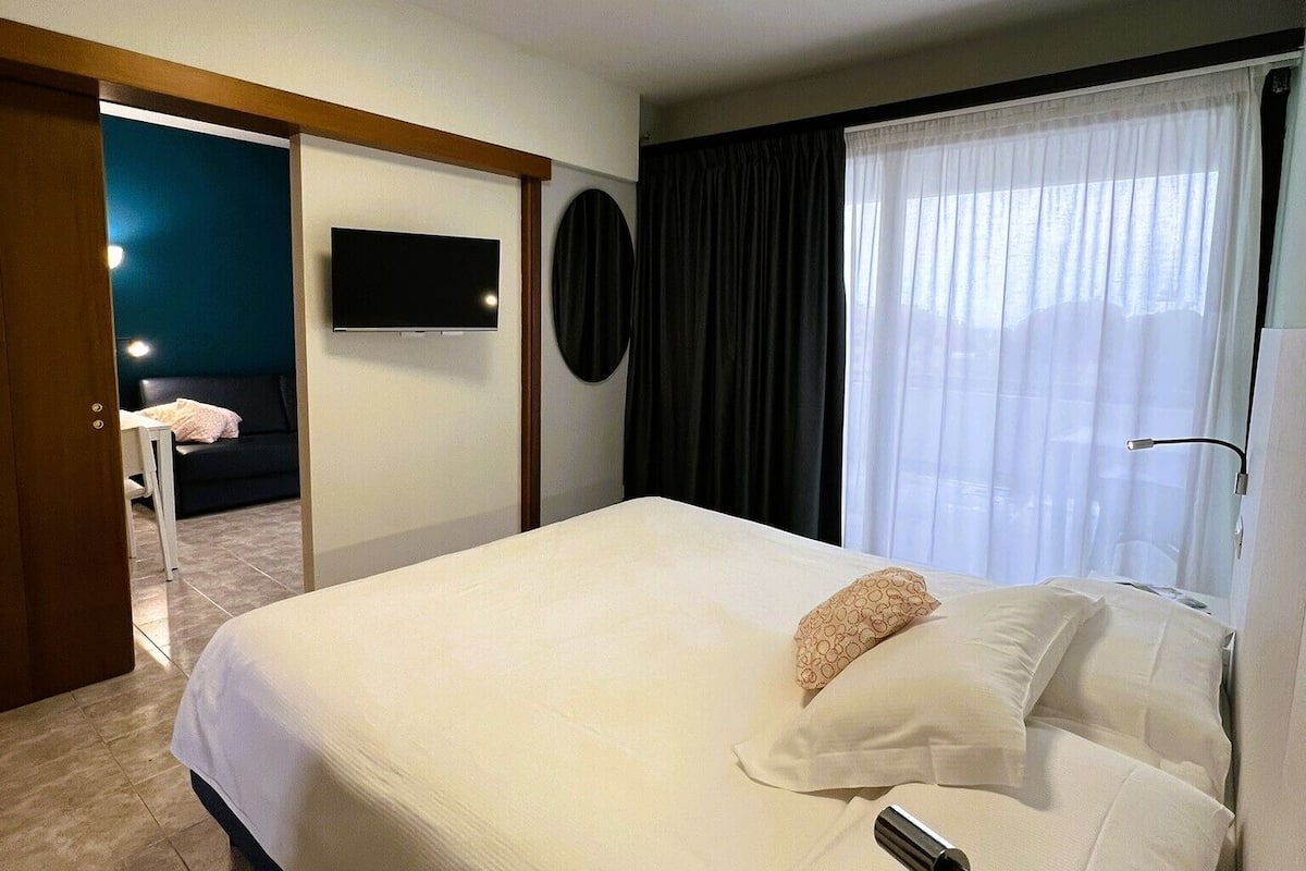 A hotel room with a bed, a TV mounted on the wall, a window with curtains, and a door leading to another room.