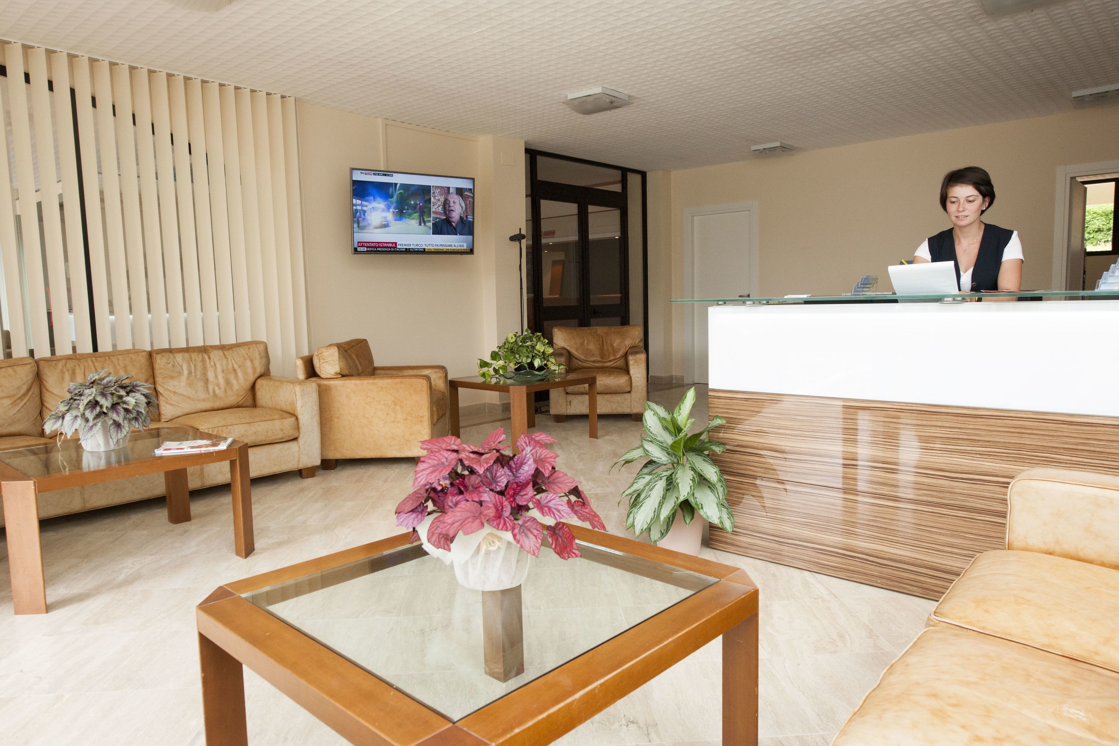 A modern reception area with a receptionist at the desk, a comfortable seating area with sofas, a glass coffee table with a vase of flowers, and a television mounted on the wall.