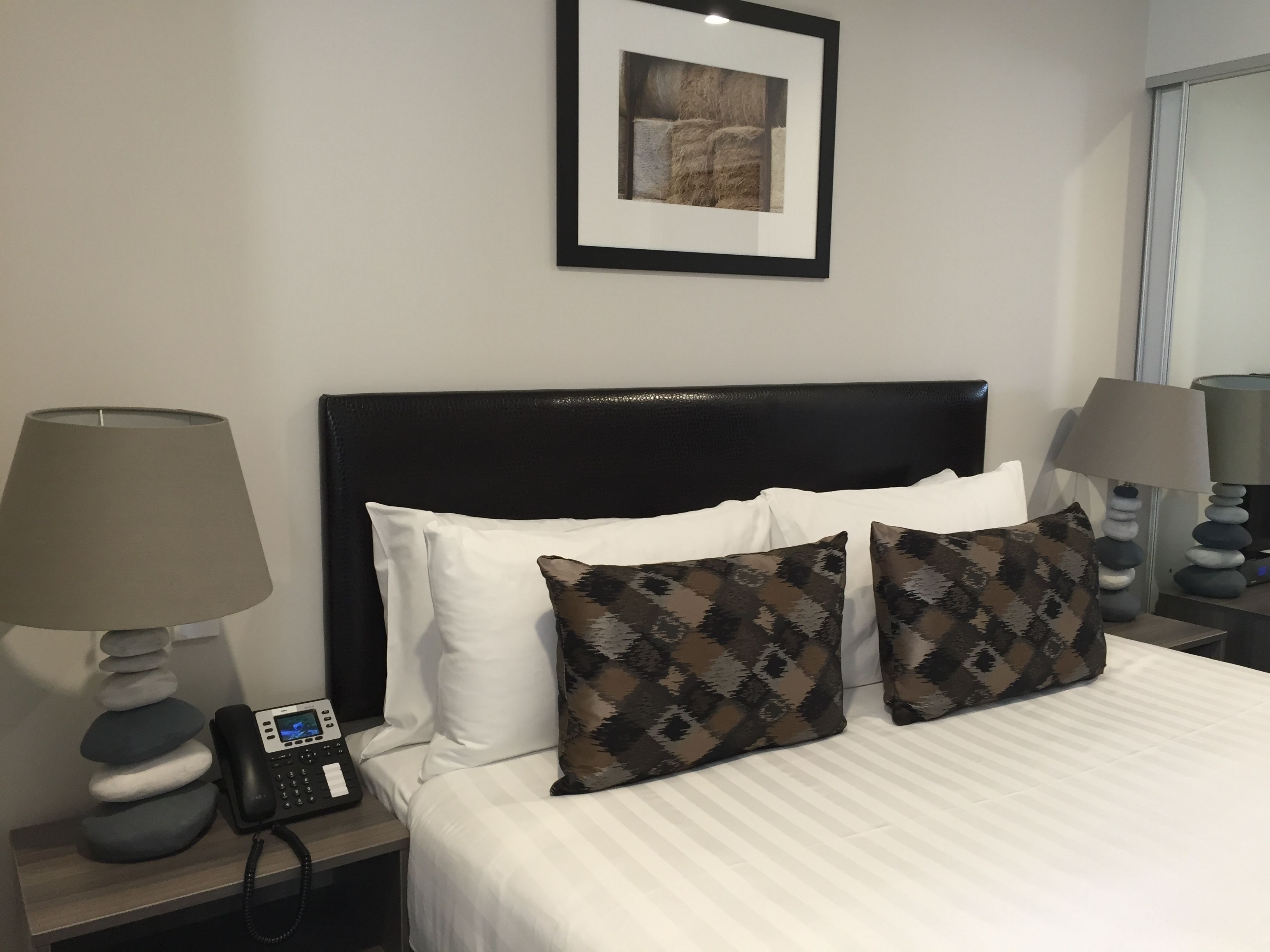 Quarto de hotel com uma cama, duas almofadas, mesa de cabeceira com abajur, telefone e quadro na parede.