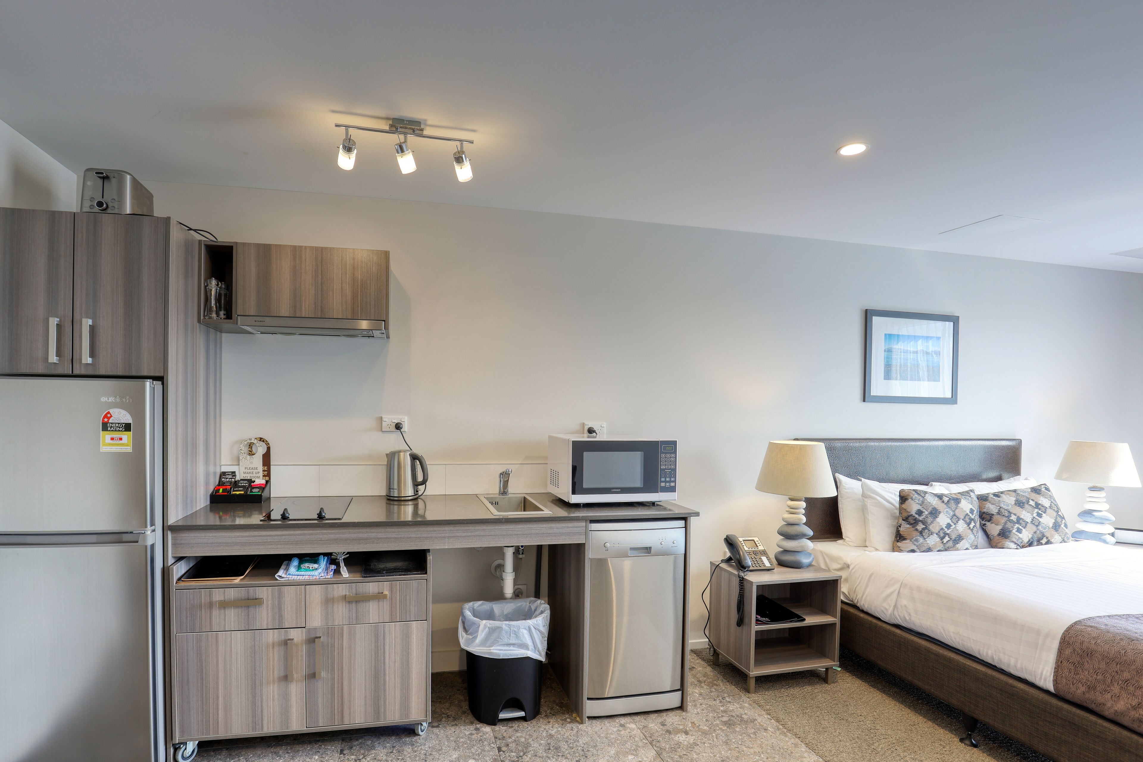 A compact hotel room with a kitchenette, a bed, bedside lamps, and a framed picture on the wall.