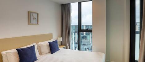 A hotel room with a large bed, a nightstand, a framed picture on the wall, and a view of the city through the window.