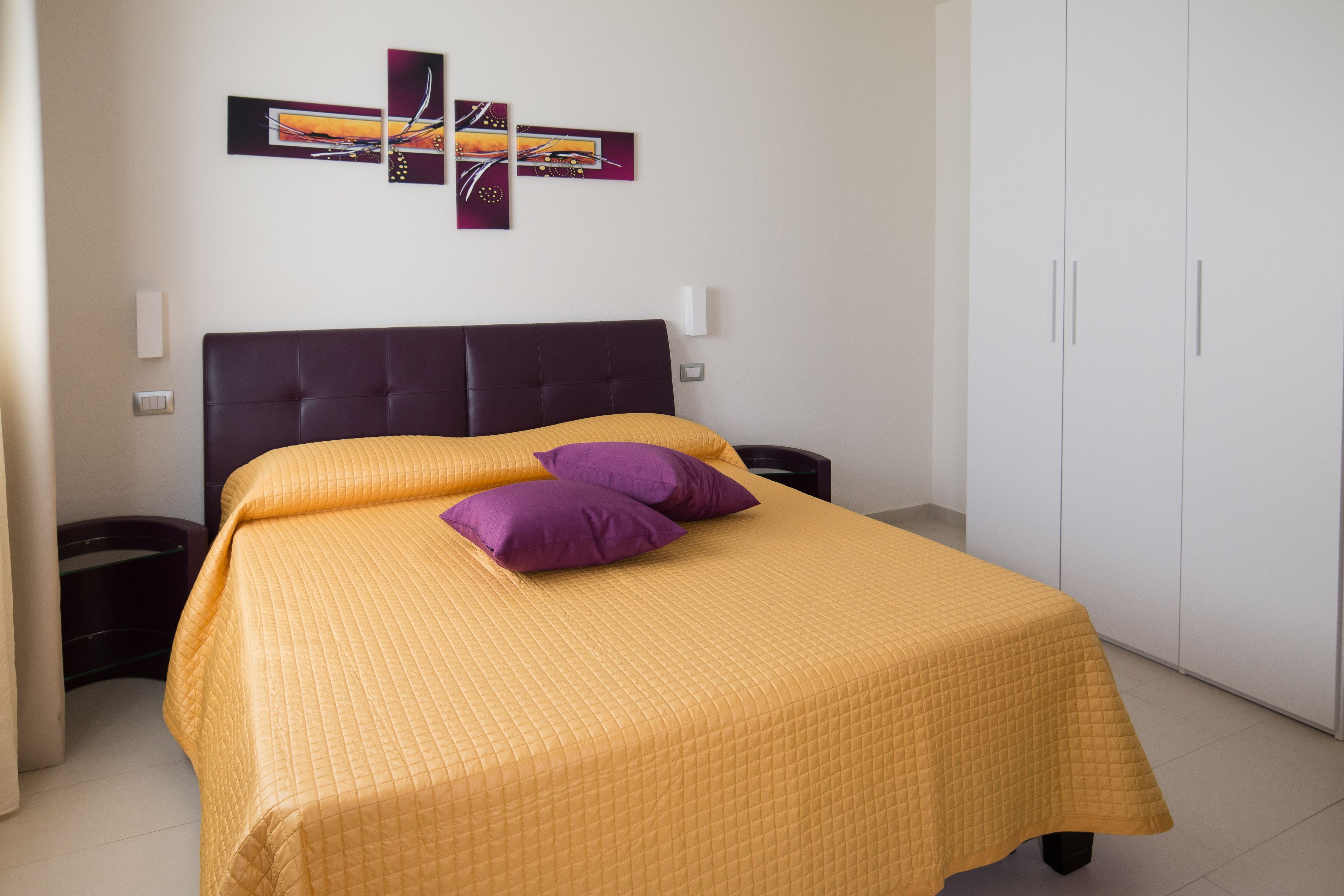 A neatly made bed with a yellow quilt and purple pillows, a dark headboard, and a white wardrobe with two doors.