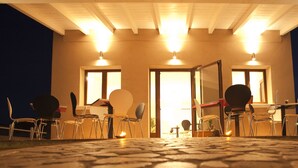 An outdoor seating area with tables and chairs, a building entrance, and a well-lit interior. - Xenia Hotel (Villasimius)