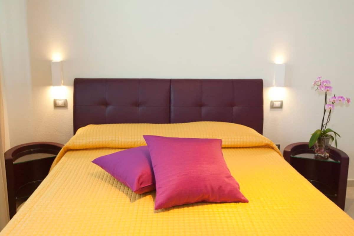 A bed with a yellow quilt and two purple pillows, flanked by nightstands with lamps and a vase of pink orchids.
