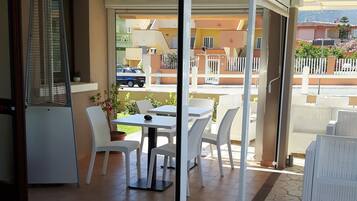 A covered outdoor area with a dining table and chairs, a refrigerator, and a potted plant.