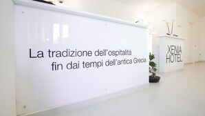 A reception area with a sign stating "La tradizione dell'ospitalità fin dai tempi dell'antica Grecia" and a "XENIA HOTEL" sign. - Xenia Hotel (Villasimius)