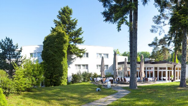 A modern building with a covered outdoor seating area, surrounded by trees and a well-maintained lawn. - Parkhotel Styria (Steyr)