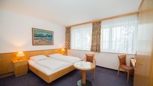A hotel room with a bed, two bedside tables with lamps, a small round table with a chair, a desk with a chair, and a window with curtains. - Parkhotel Styria (Steyr)