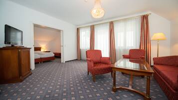 A hotel room with a bed, two red armchairs, a glass-top coffee table, a television on a wooden cabinet, and a chandelier.