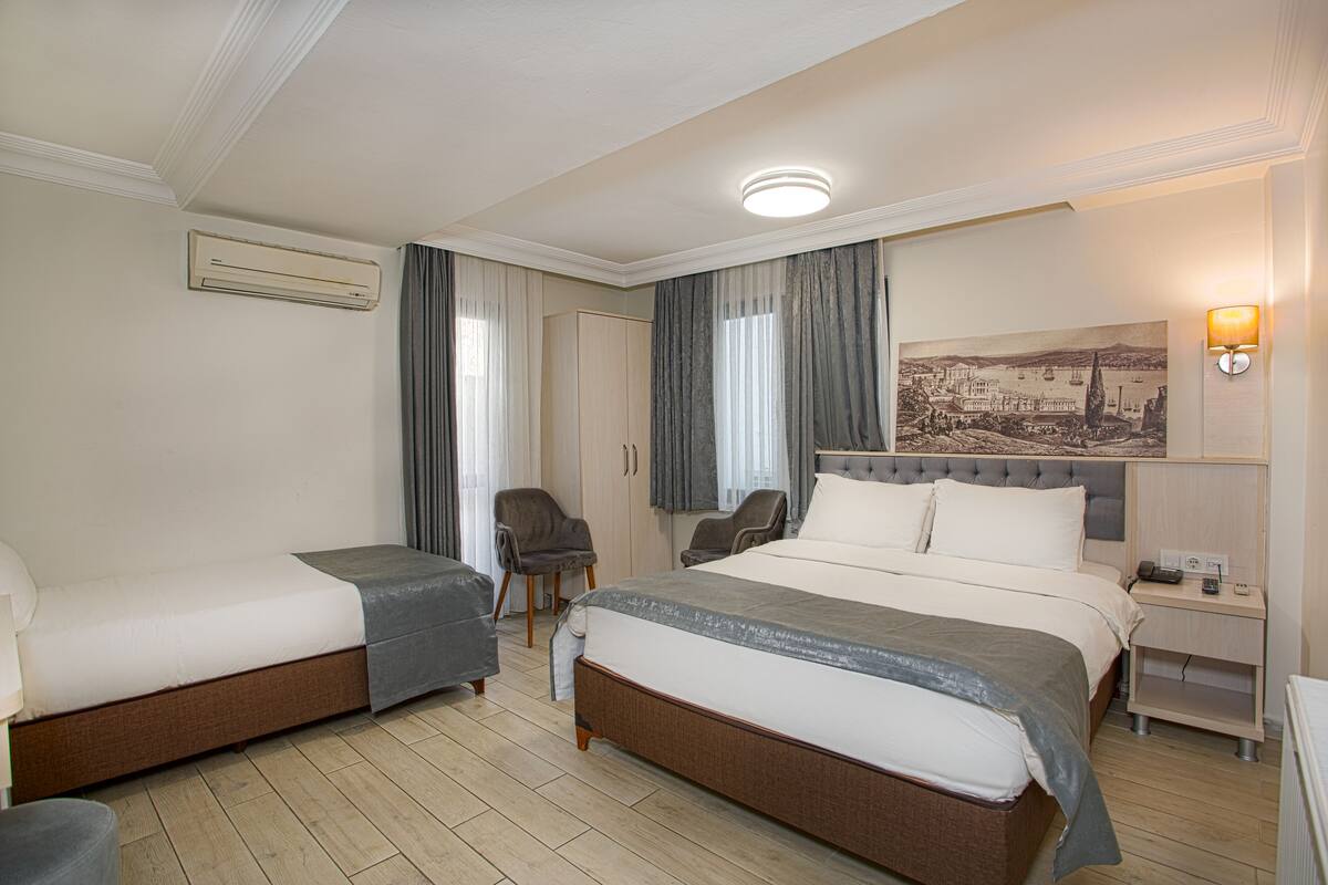 A hotel room with two single beds, a sitting area with a chair, a desk with a lamp, and a wall-mounted air conditioner.