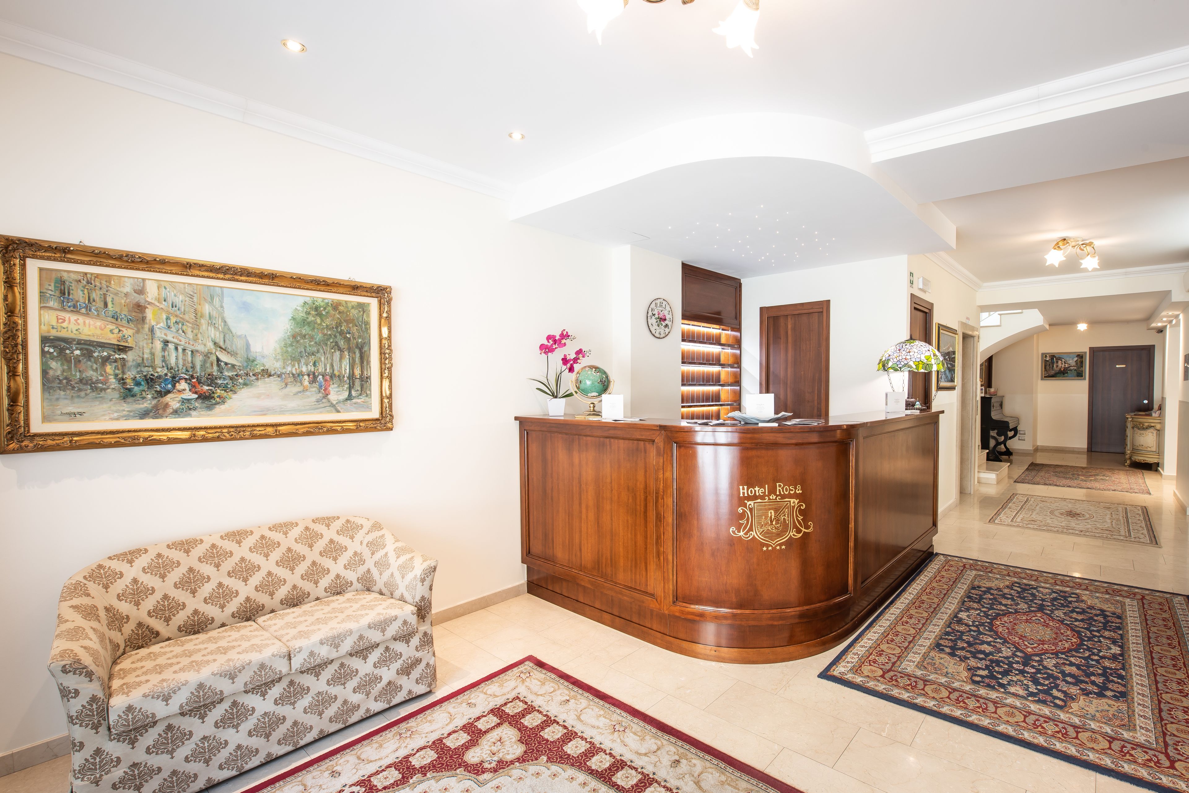 Un'area reception di un hotel con una scrivania in legno, un divano, un quadro di grandi dimensioni e un tappeto decorato.
