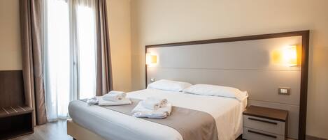 A hotel room with a large bed, bedside tables, a bench, and a window with curtains.