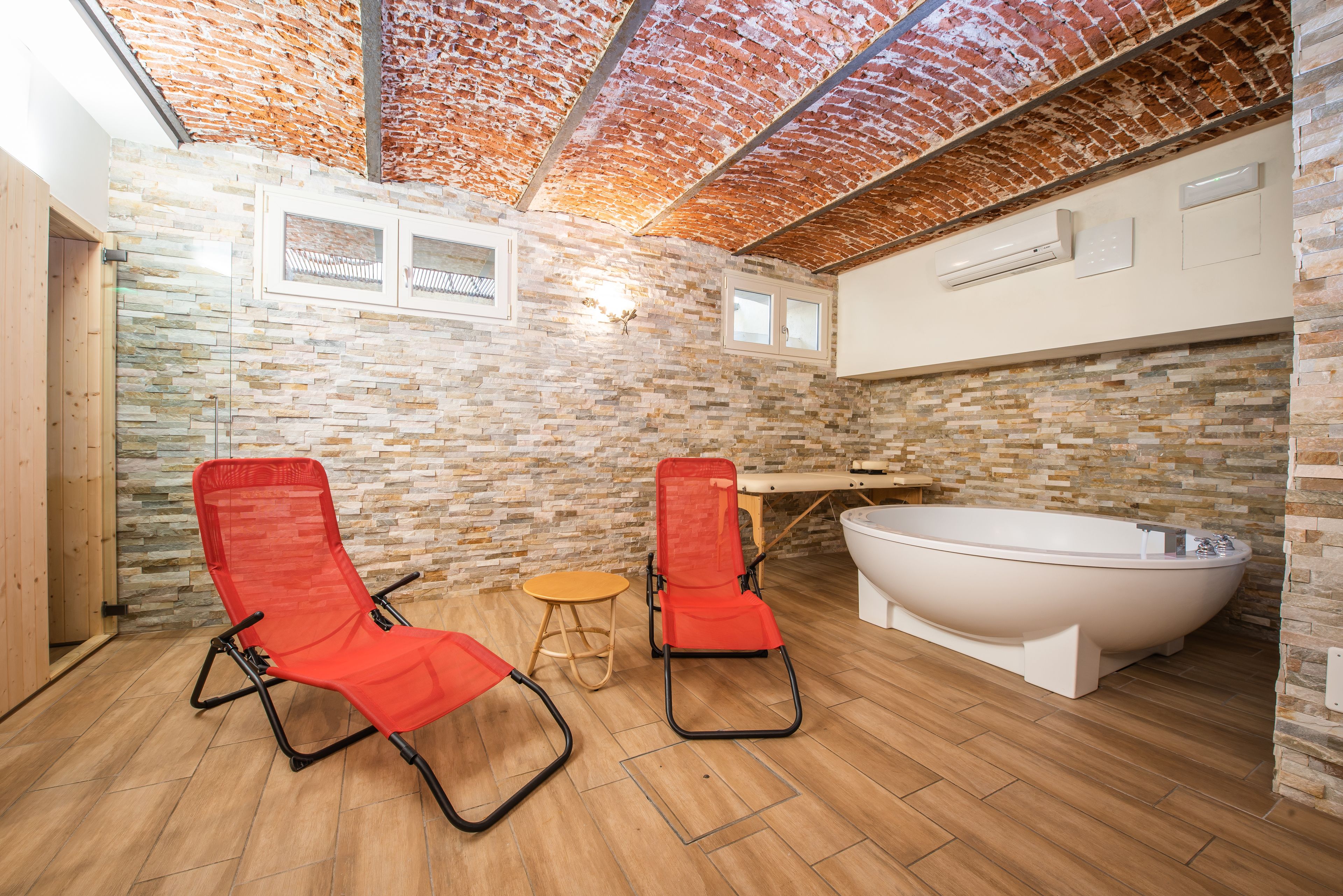 A bathroom with a stone wall, two red chairs, a bathtub, and a wooden floor.