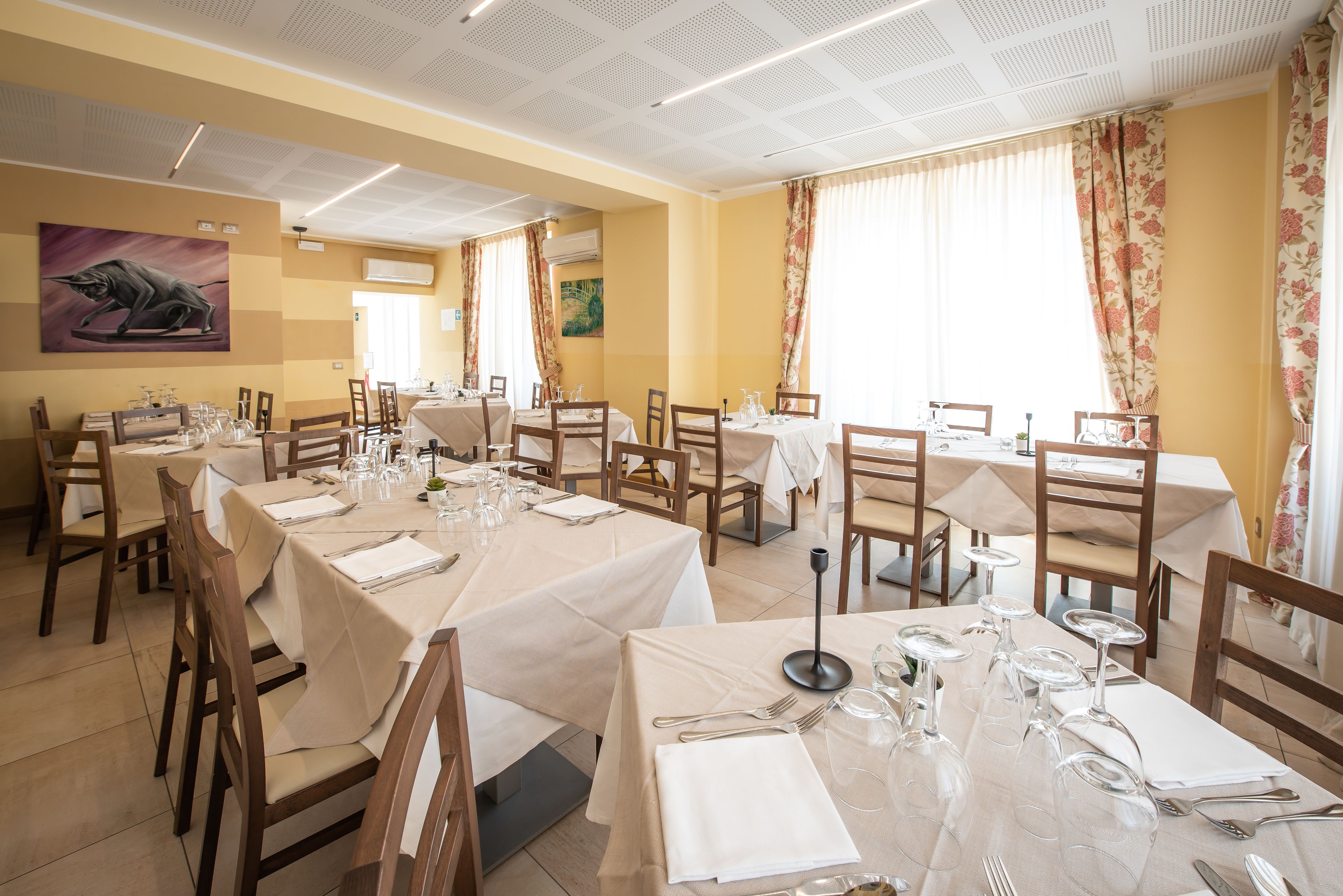 Un restaurant doté de nombreuses tables dressées pour le service, avec un plafond orné d’un motif en réseau et des rideaux à motifs floraux.
