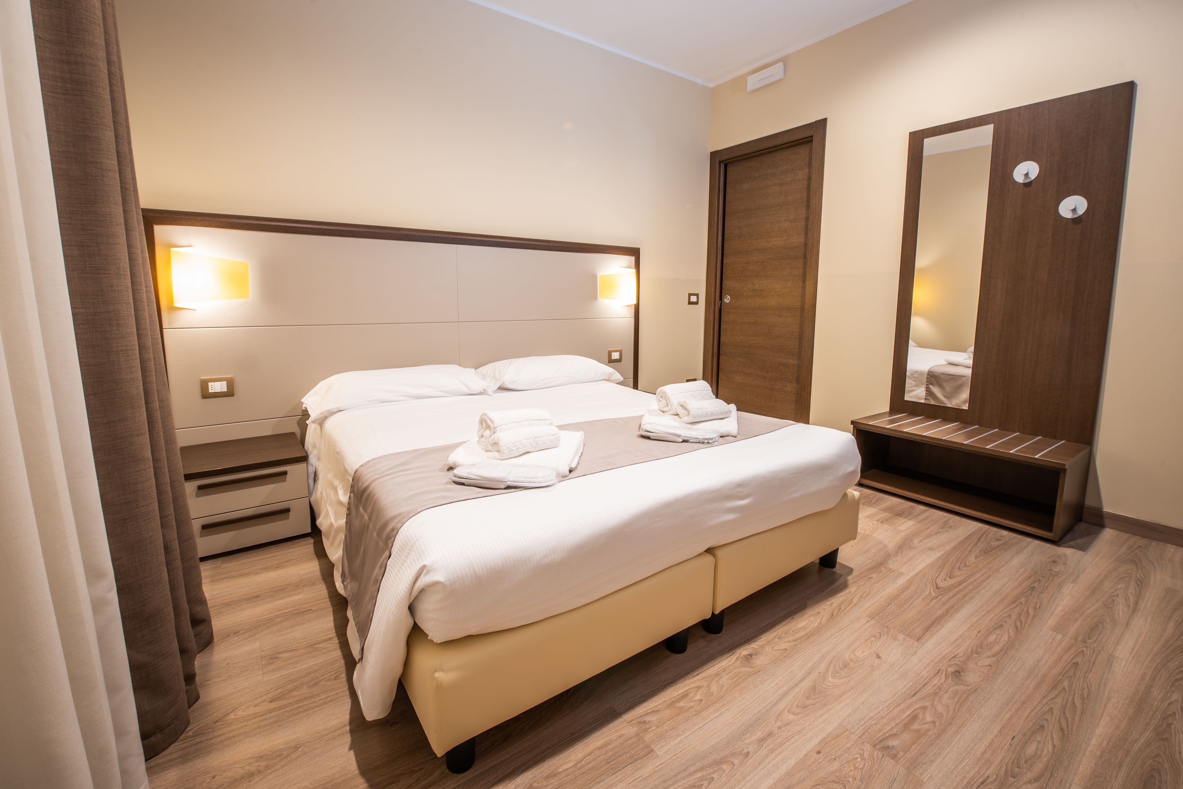 Habitación de hotel con una cama grande, lámparas de noche, suelo de madera, armario y espejo.