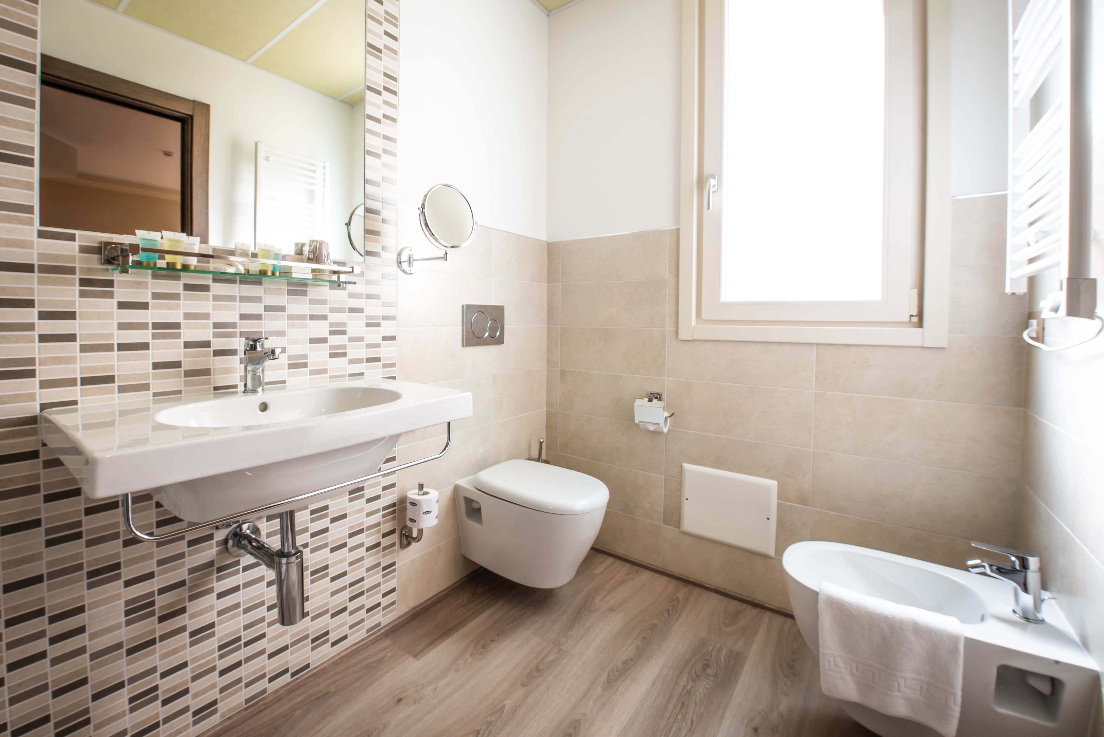 Une salle de bain moderne avec un lavabo, des toilettes et une baignoire blancs, un miroir et une fenêtre laissant entrer la lumière naturelle.