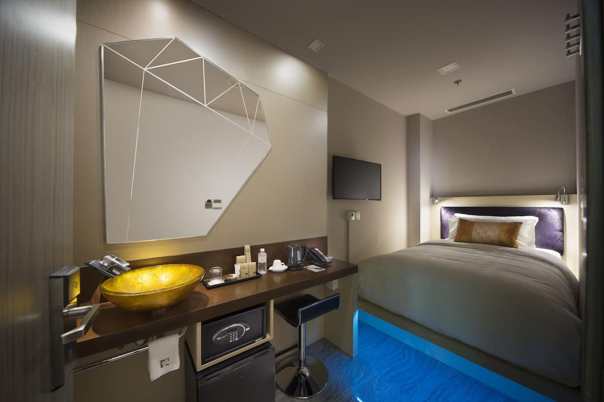 A modern hotel room with a large mirror, a gold bowl sink, a flat-screen TV, a bed with a headboard, and a small desk with a coffee maker.