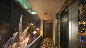 Un pasillo moderno con piso estampado, un muro de cristal con iluminación decorativa y un ascensor con pantalla digital.