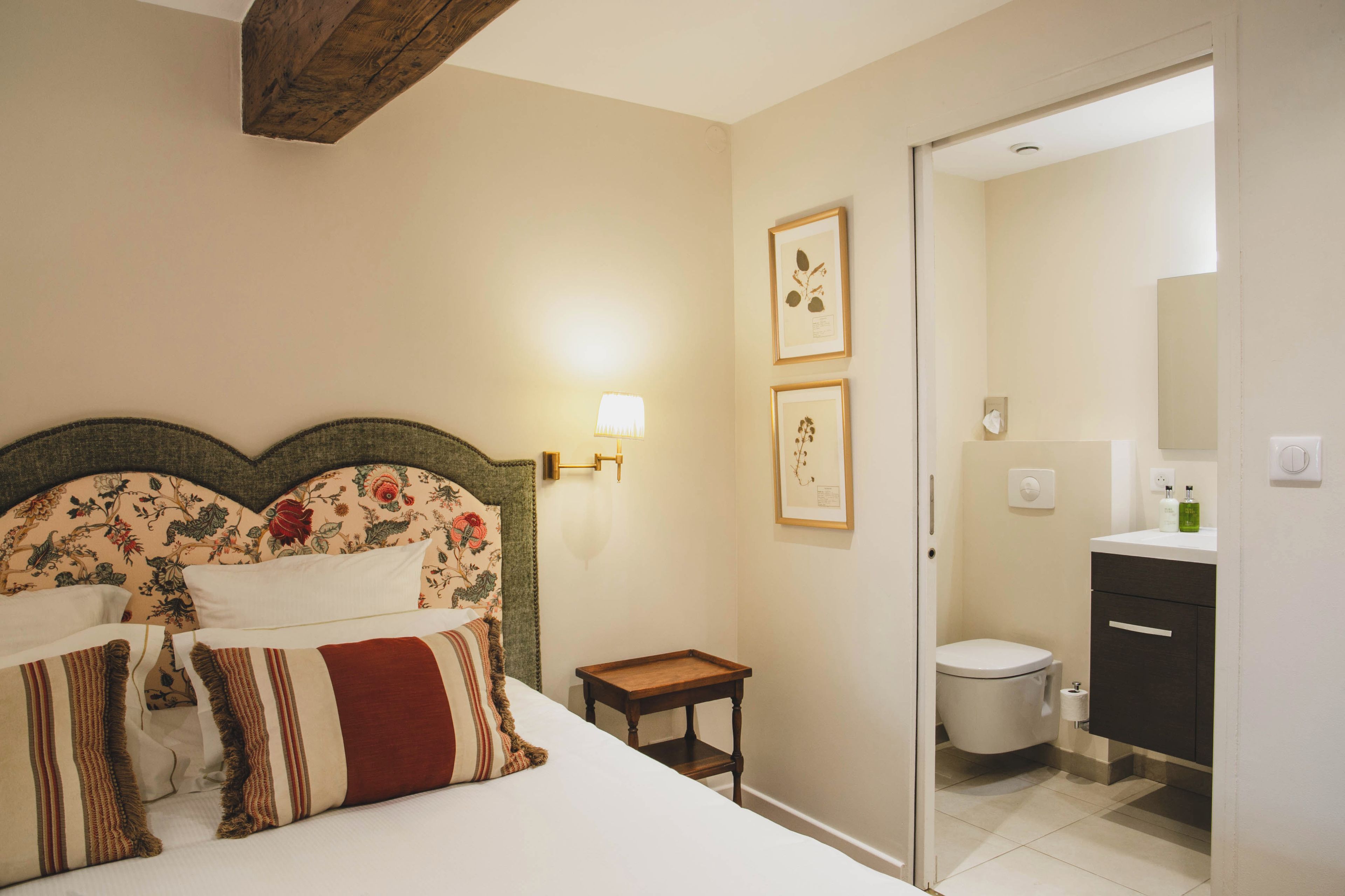 Un dormitorio con cama, mesita de noche, lámpara de pared e impresiones botánicas enmarcadas.