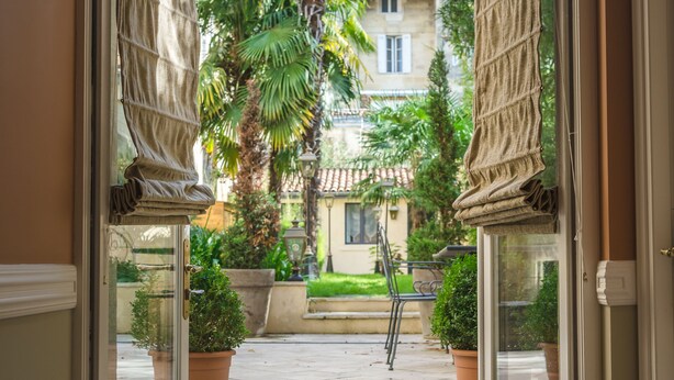 An open door leading to a courtyard with potted plants, a palm tree, and a building in the background. - Villa Victor Louis (Bordeaux)