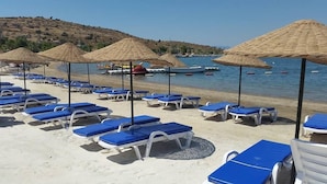 Une plage bordée de rangées de chaises longues bleues et de parasols en paille, une rive sablonneuse et des bateaux sur l’eau.