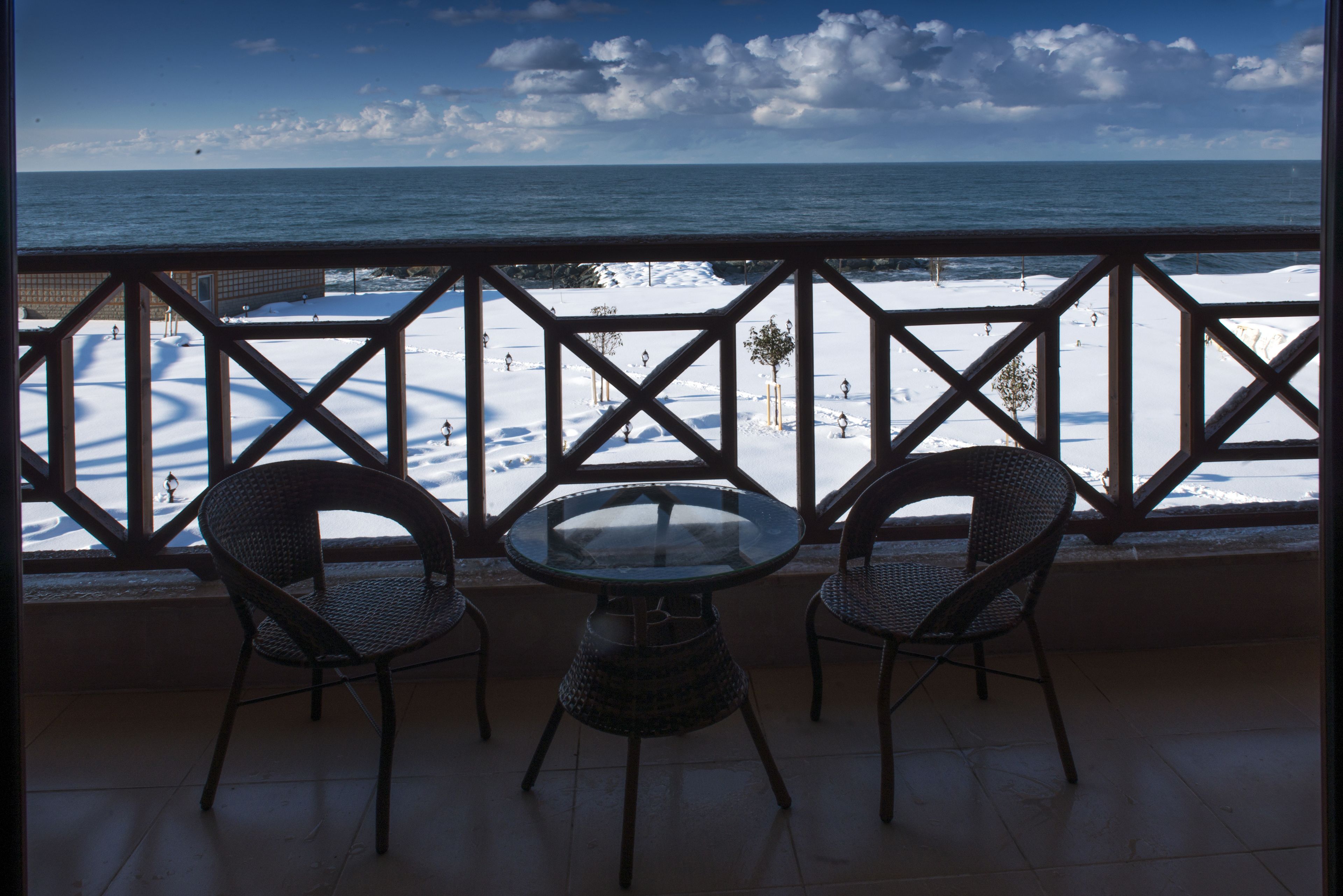 A balcony with a glass table and two wicker chairs, overlooking a snowy beach and the ocean.