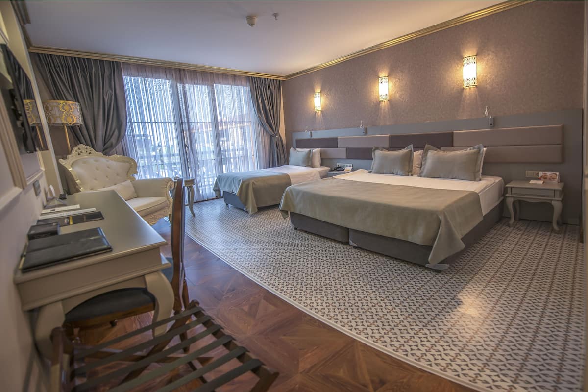 Une chambre d’hôtel avec un grand lit, deux tables de chevet, un bureau et une chaise. On y trouve des rideaux, des appliques murales et un tapis à motifs.