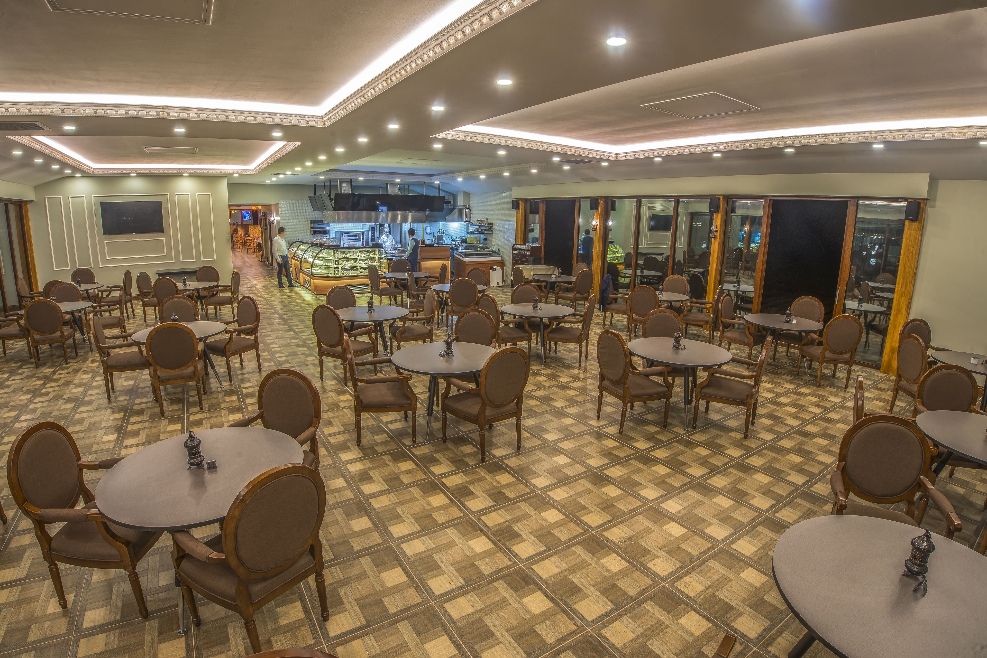 A spacious dining area with multiple round tables and chairs, a central kitchen area, and a view of the cityscape through large windows.