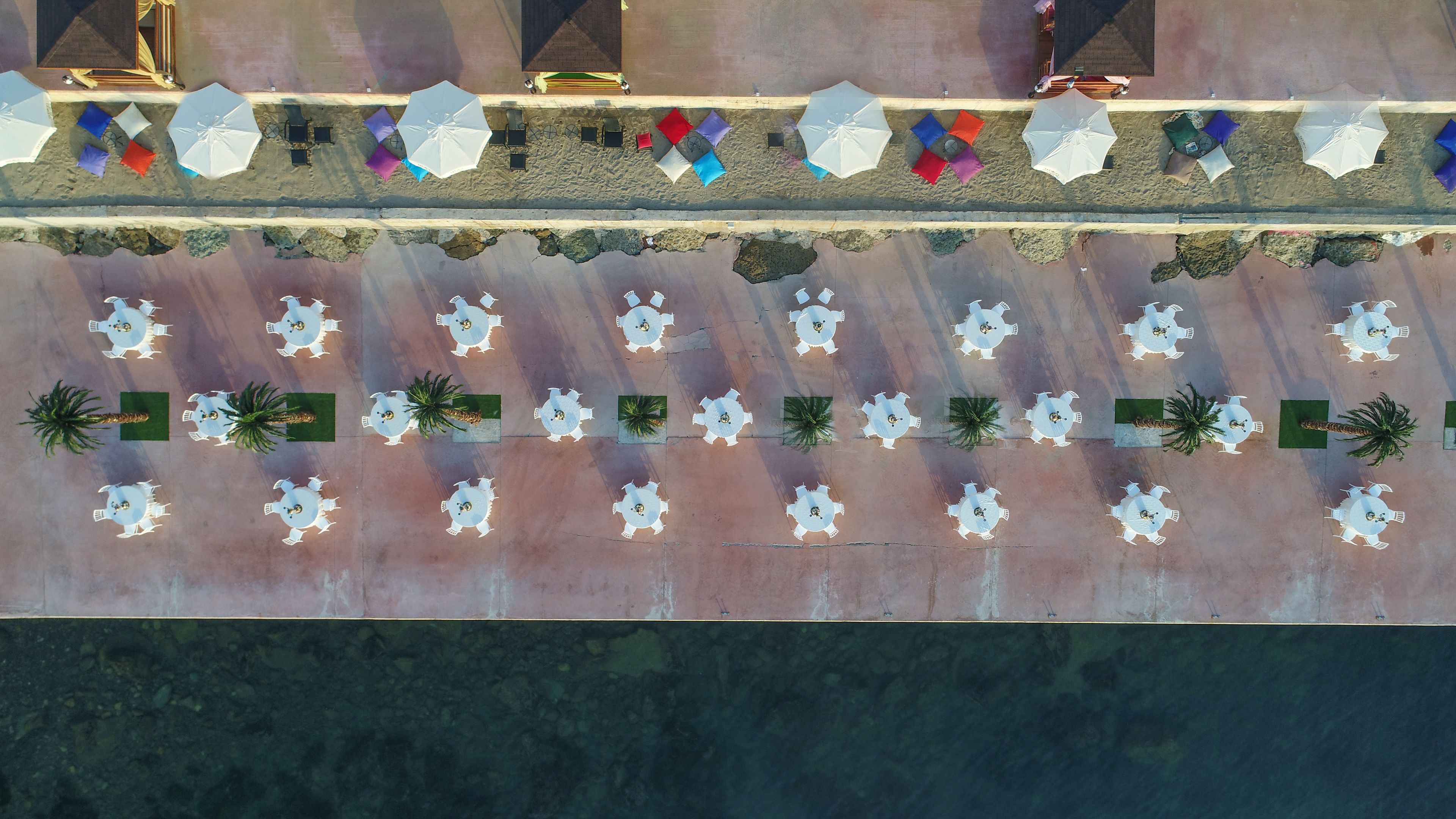 An aerial view of a beachside resort with multiple dining areas, each with tables and chairs, and palm trees.