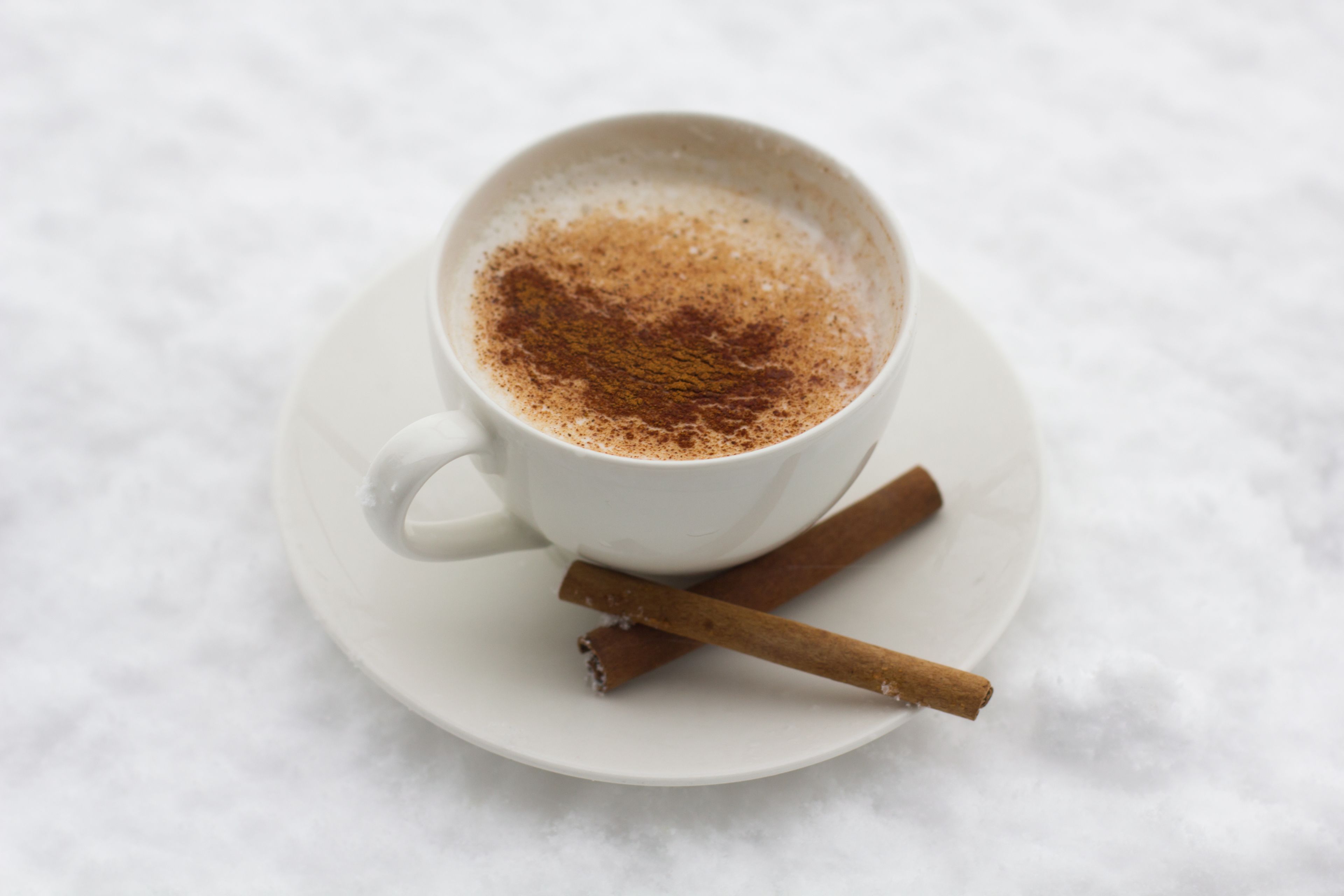Een witte koffiekop met kaneel op een schotel, vergezeld van een kaneelstokje.