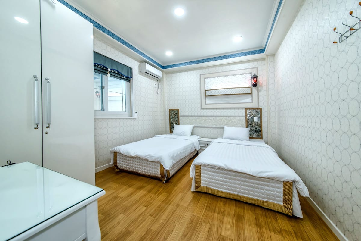 A bedroom with two beds, a glass table, wooden flooring, and a window with blinds.