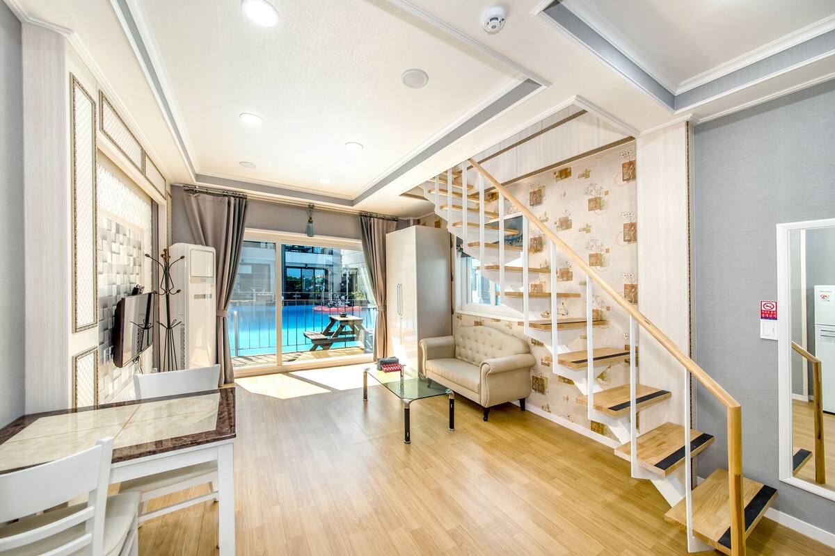A spacious living area with a wooden floor, a glass coffee table, a sofa, and a staircase with wooden railings.