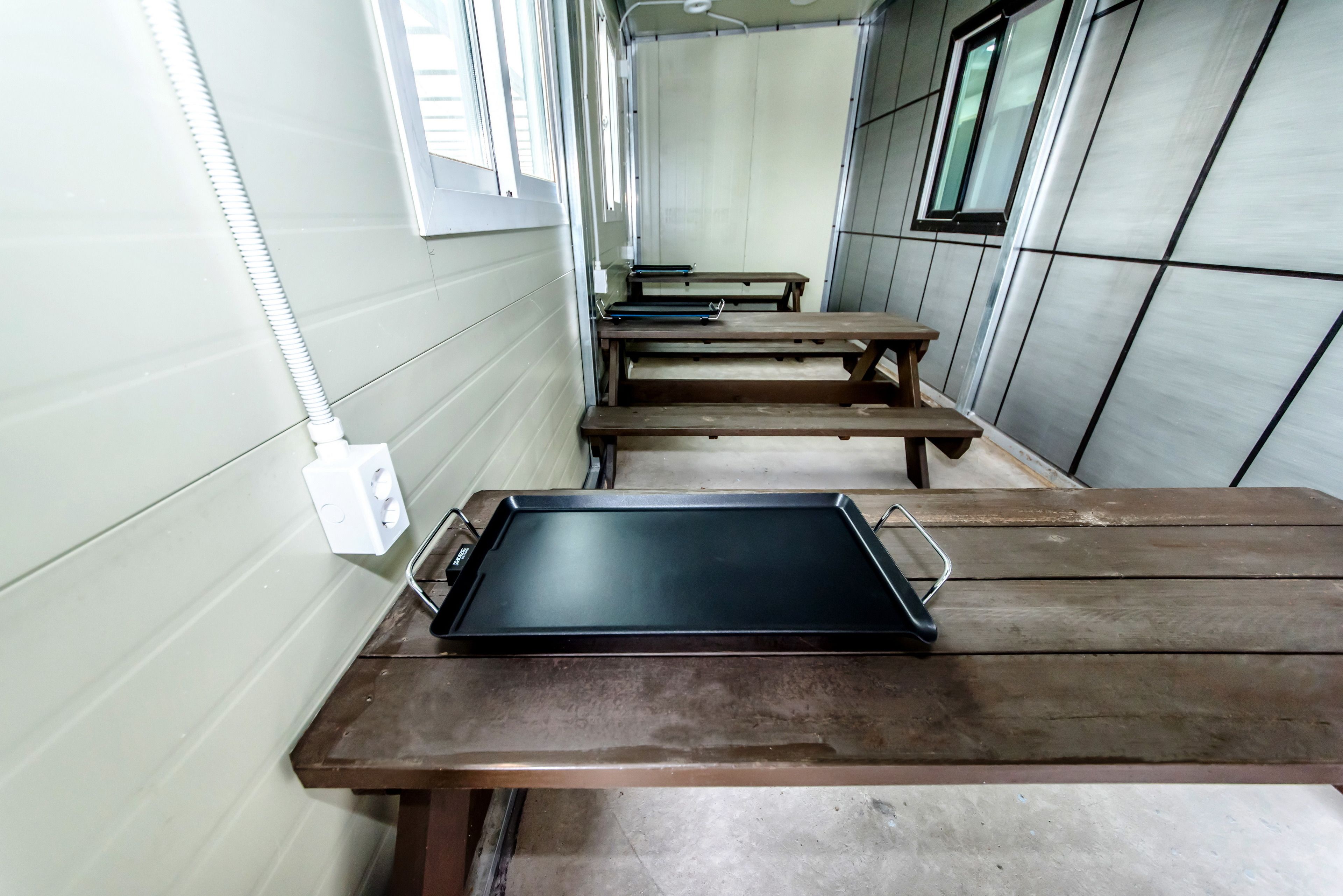 A row of wooden tables with a black tray on one, a white electrical outlet on the wall, and a window with blinds.